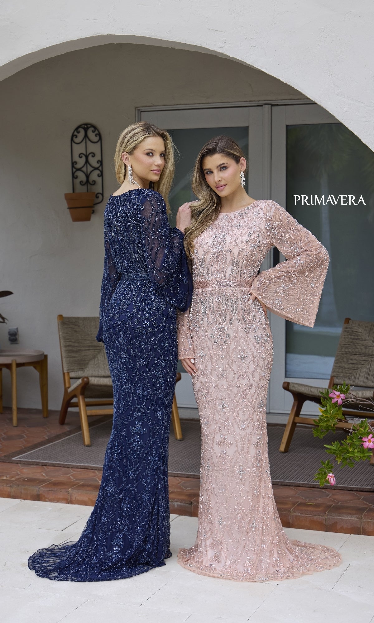 Two women pose outdoors in floor-length Primavera 12187 formal dresses—one in navy blue with long bell sleeves, the other in light pink—in front of a patio.